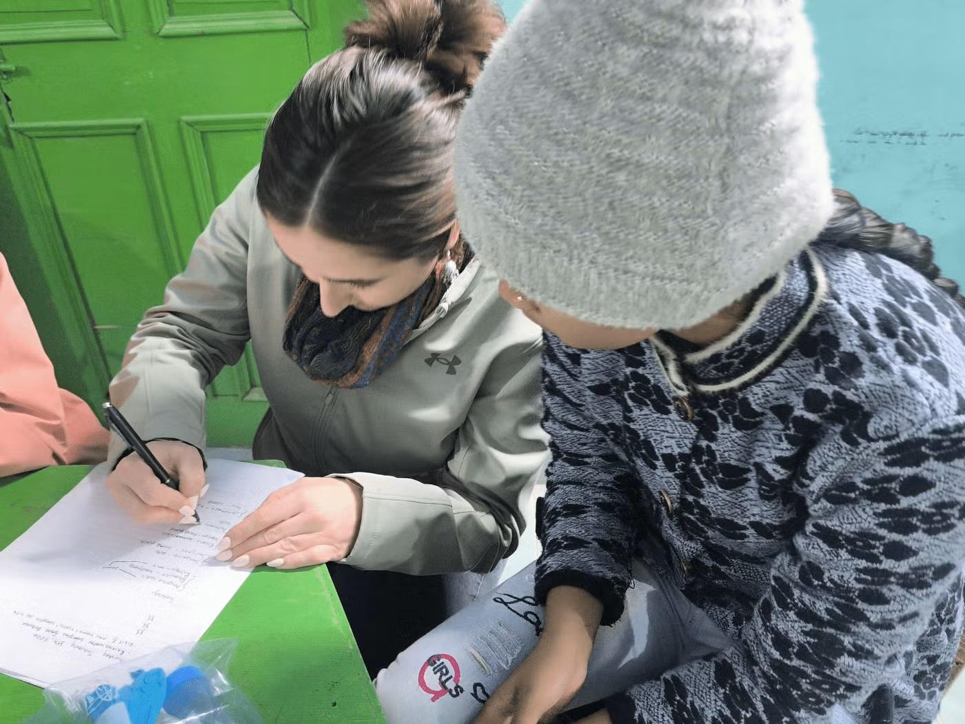 Two students lean over a green table, the one on the left is writing on a worksheet
