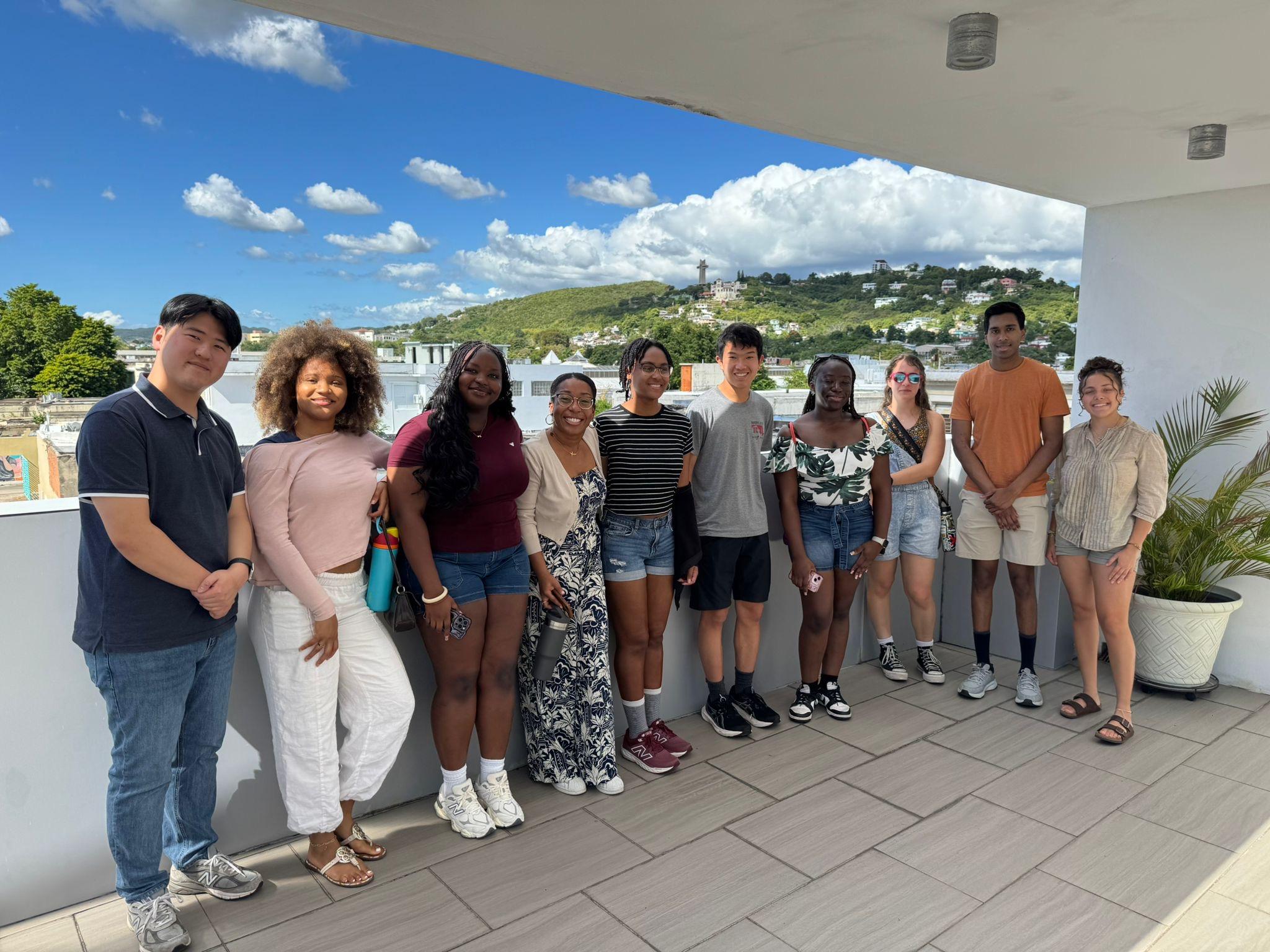 Daniel Lee (left) stands with a group of students and other program leaders on an outdoor rooftop terrace in Ponce, Puerto Rico, smiling at the camera. A hillside with green trees and buildings is visible in the background under a bright blue sky with white clouds. 