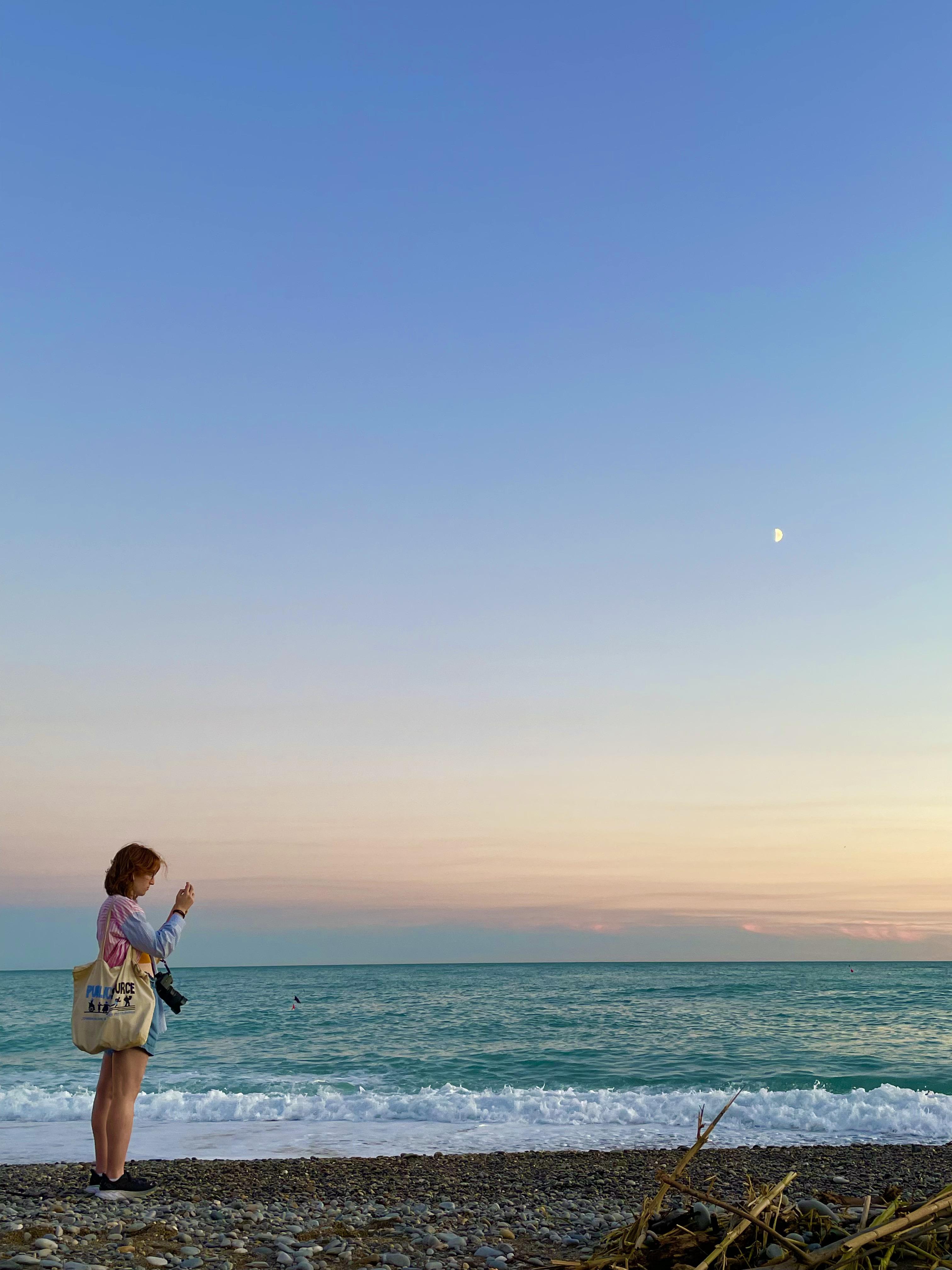 Jess stands on a rocky beach at sunset, taking a photo of the ocean as gentle waves roll in beneath a pastel sky with a faint moon.