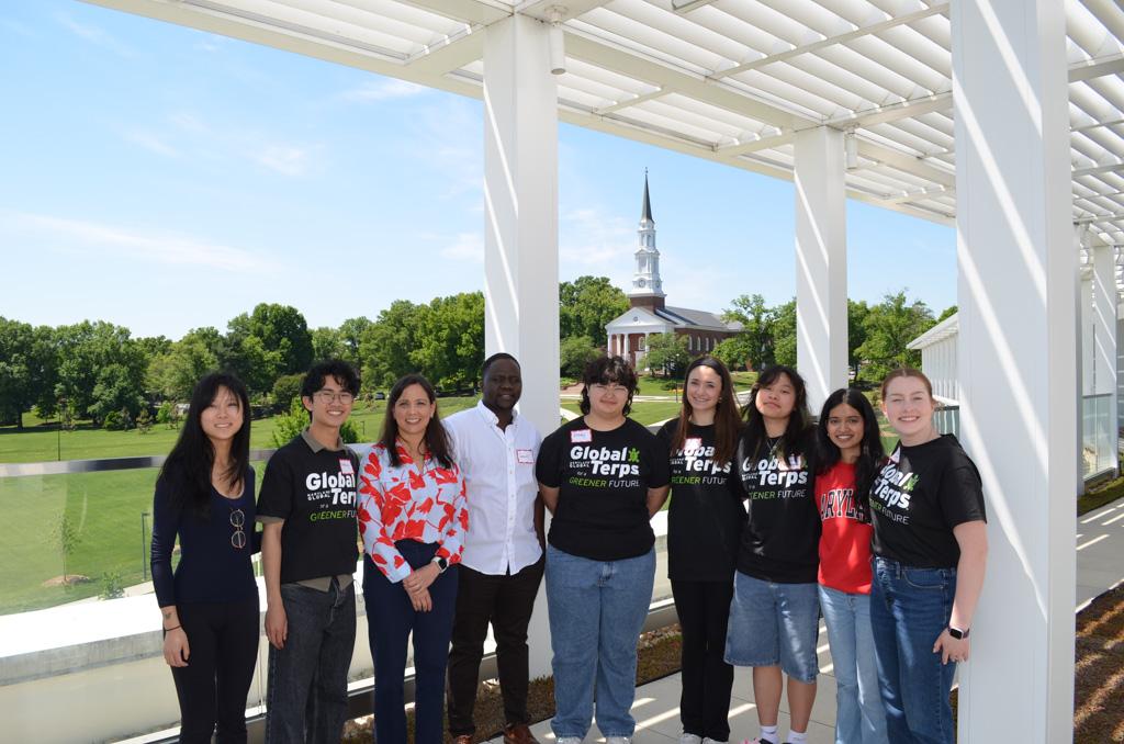 Nine individuals, some wearing Global Terps for a Greener Future, stand together on a rooftop with the UMD Chapel in the background