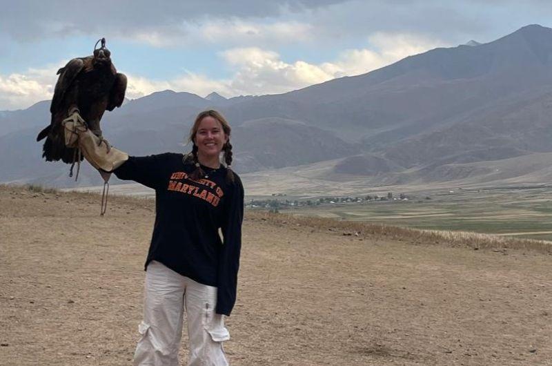 Elena stands in a wide, open landscape with mountains in the background under a cloudy sky, holding a large bird of prey perched on a gloved hand. The student is wearing a University of Maryland shirt and light-colored pants. 