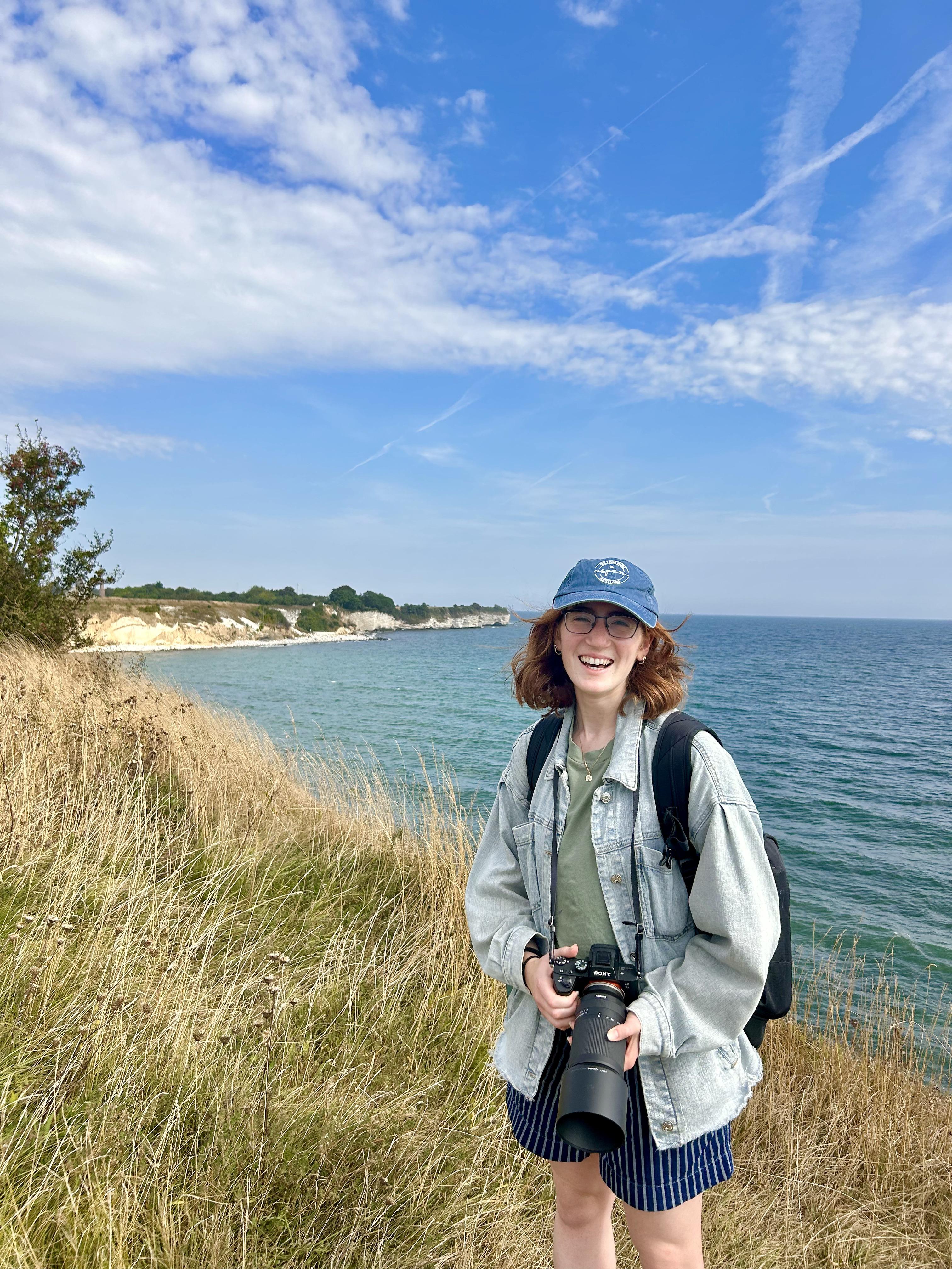 Jess stands on a grassy coastal cliff holding a camera, smiling with the ocean and white cliffs in the background under a bright blue sky.