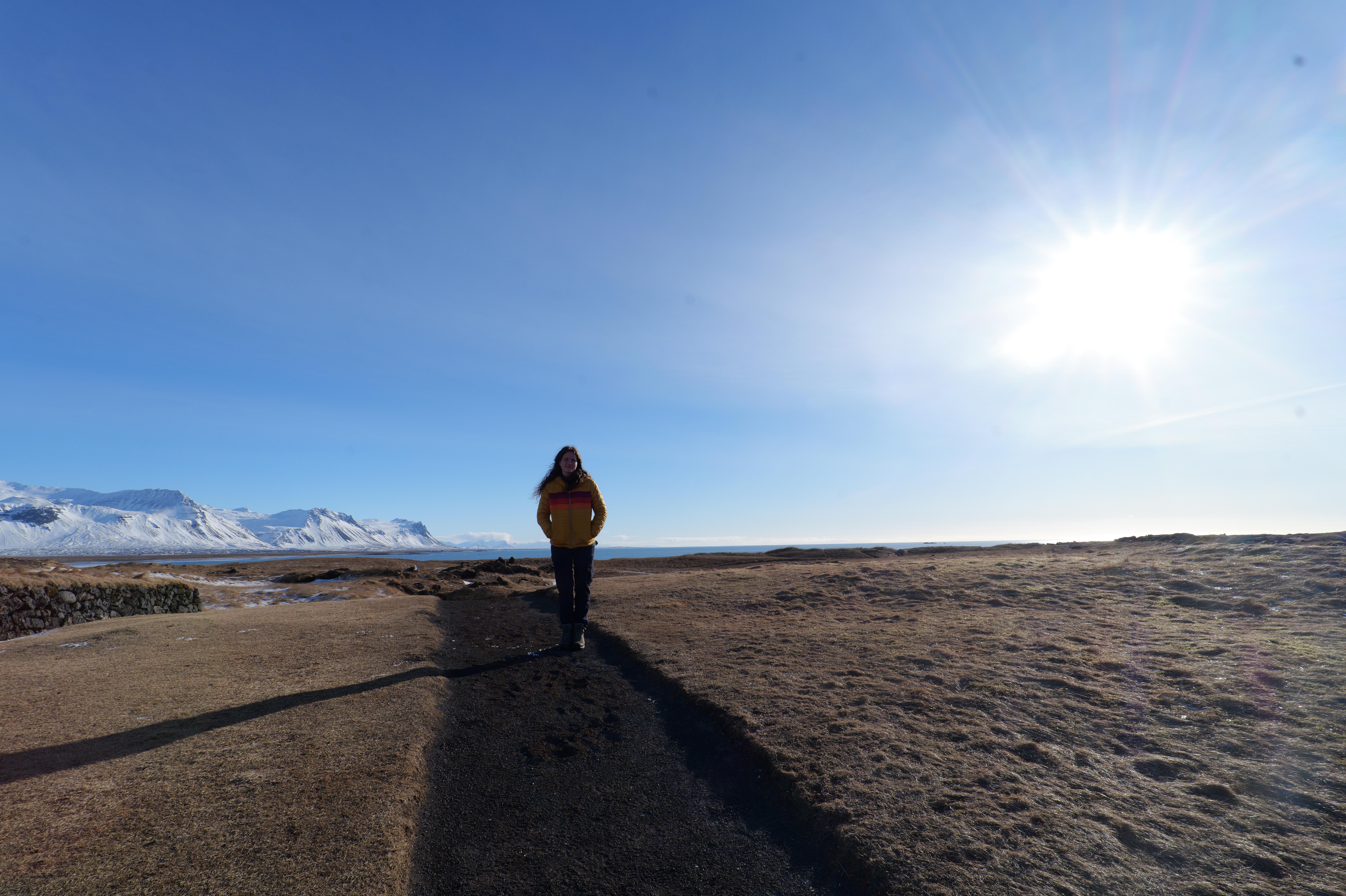 A student stands on a dirt path in a wide open Icelandic landscape with snow-covered mountains and a body of water in the distance under a bright, sunny sky.