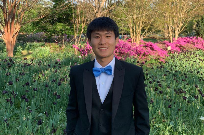 Yipeng stands in front of greenery and flowers wearing a suit and bowtie.