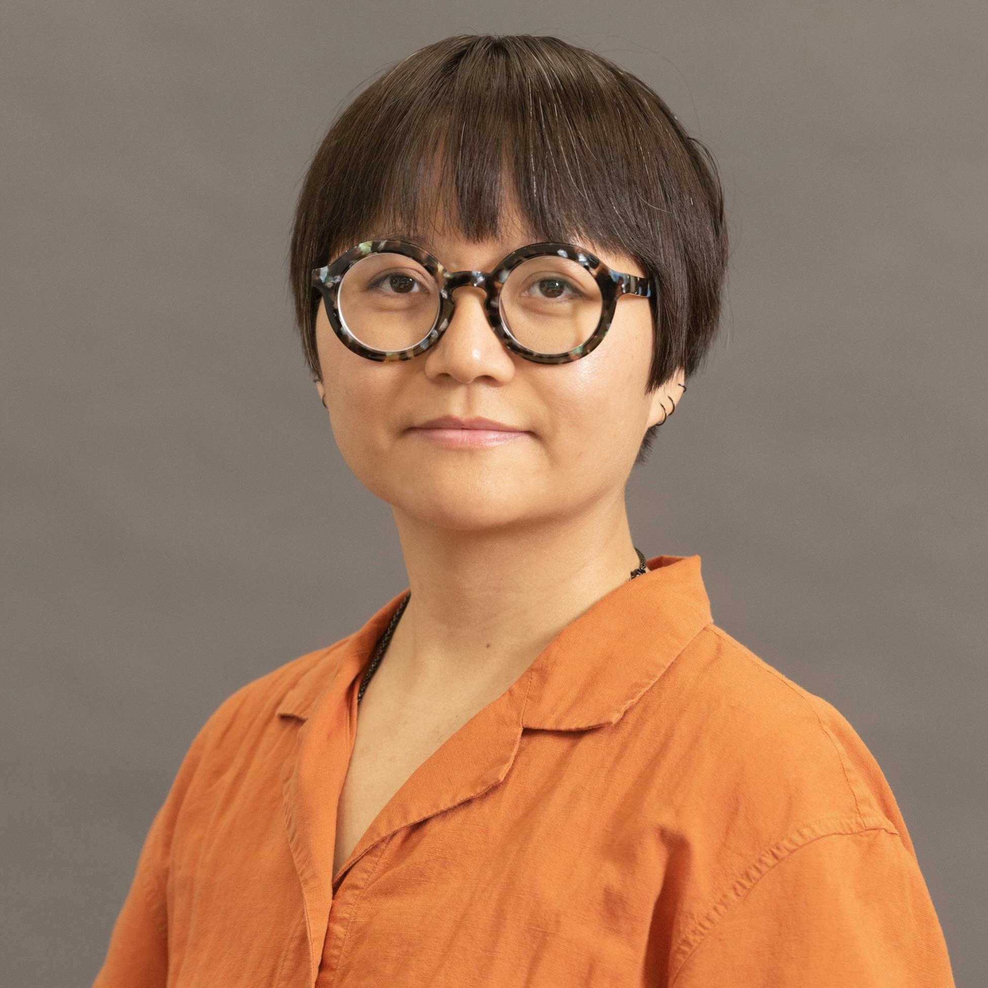 Headshot of Joan. She has short brown hair, round glasses and wears an orange button down shirt.