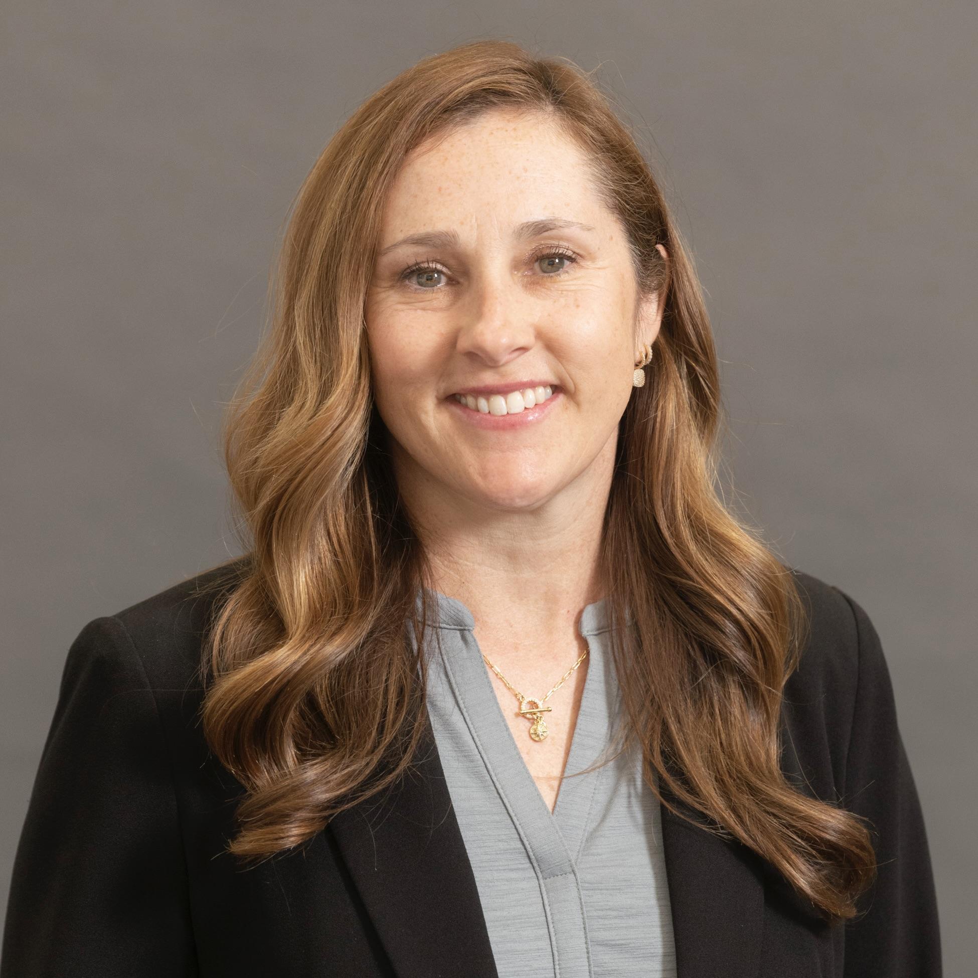Professional headshot of Jamie. She has long brown hair and wears a dark blazer with grey top.
