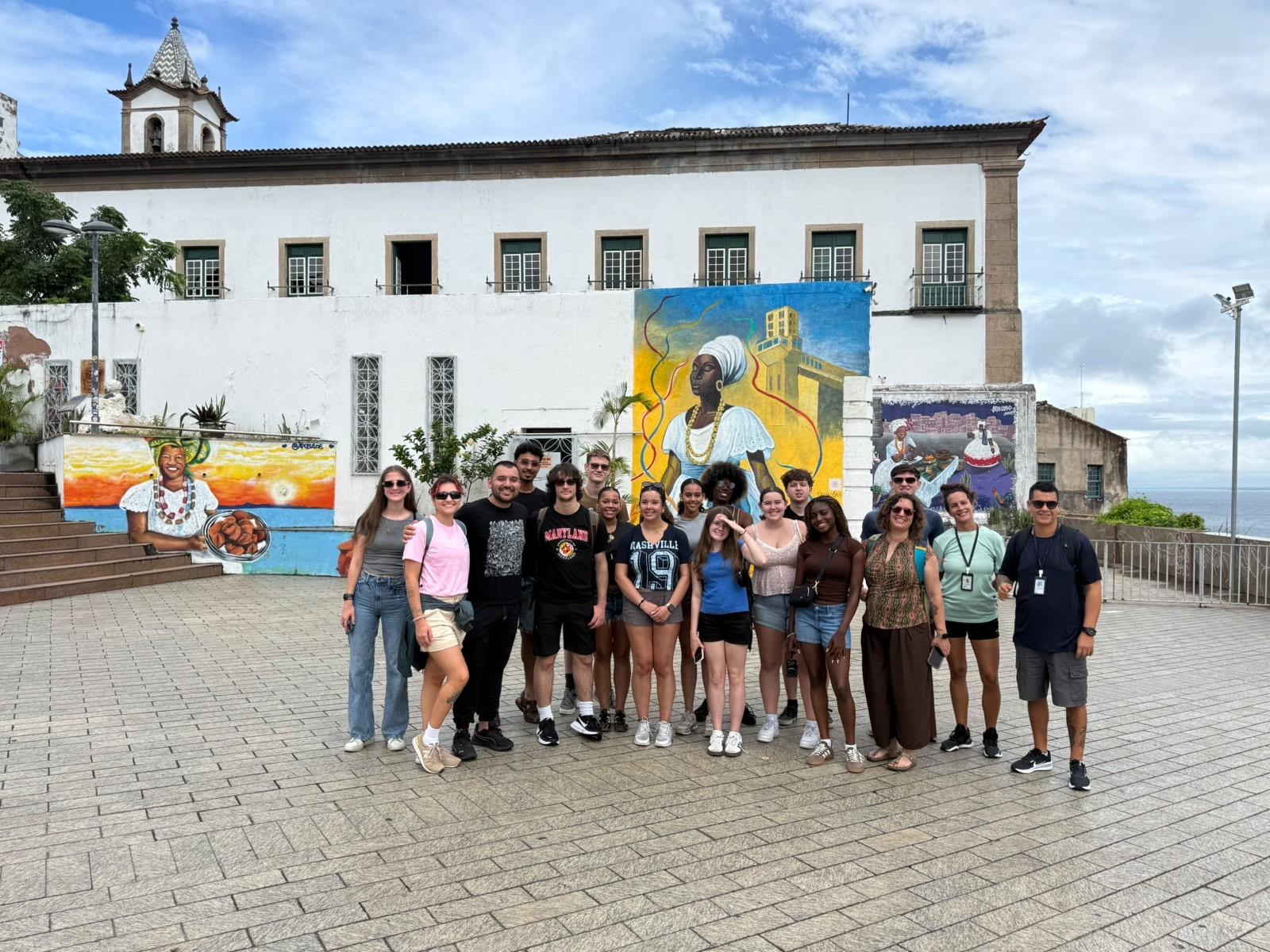 A group of students and faculty pose together in a plaza in front of a historic white building decorated with colorful murals.